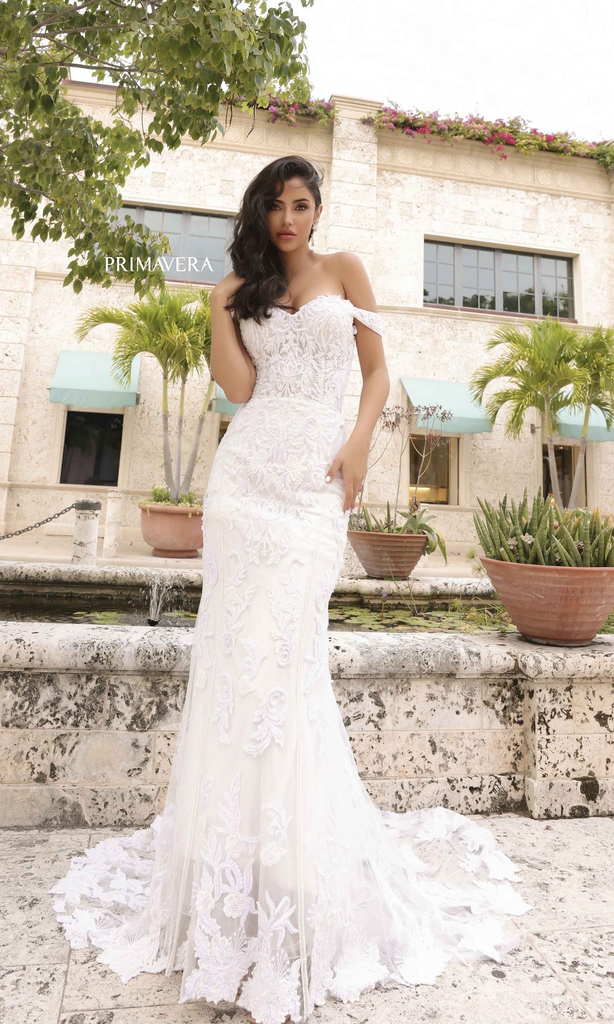 A woman models the Long Wedding Dress 11104 by Primavera with lace embroidery and a flowing train, posing outdoors before a stone building with large windows, potted plants, and palm trees.